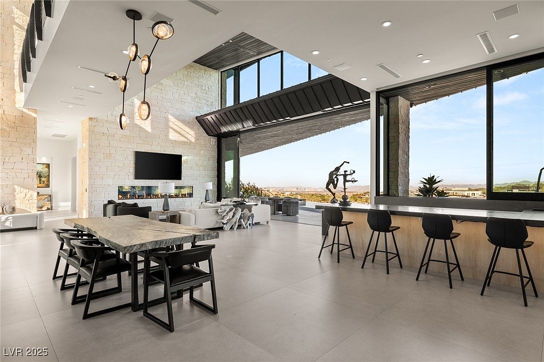 This is a spacious and modern living room featuring a large dining table with black chairs, a sleek fireplace, and a comfortable seating area. The room boasts high ceilings, large windows offering scenic views, and a stylish bar area with stools. The open floor plan and contemporary design create an inviting and luxurious atmosphere.