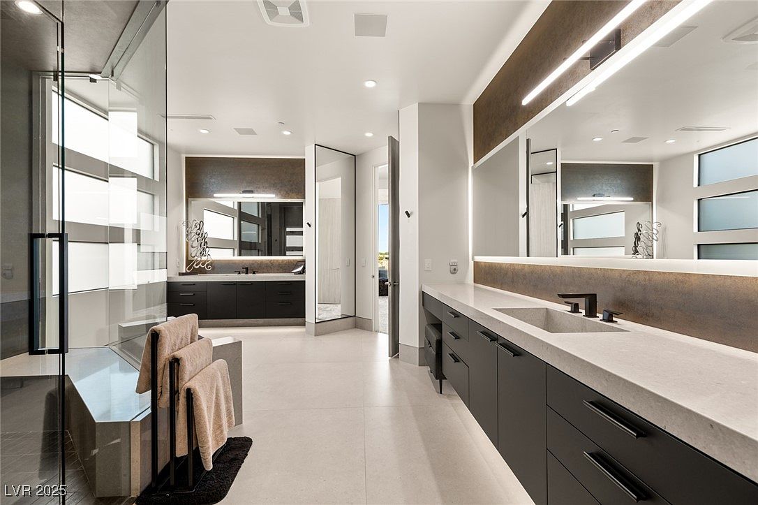 This is a modern primary bathroom featuring a large walk-in shower with glass enclosure, dual vanities with dark cabinetry and light countertops, and a neutral color palette. The bathroom is spacious and well-lit, with clean lines and a minimalist design. The overall impression is luxurious and serene.