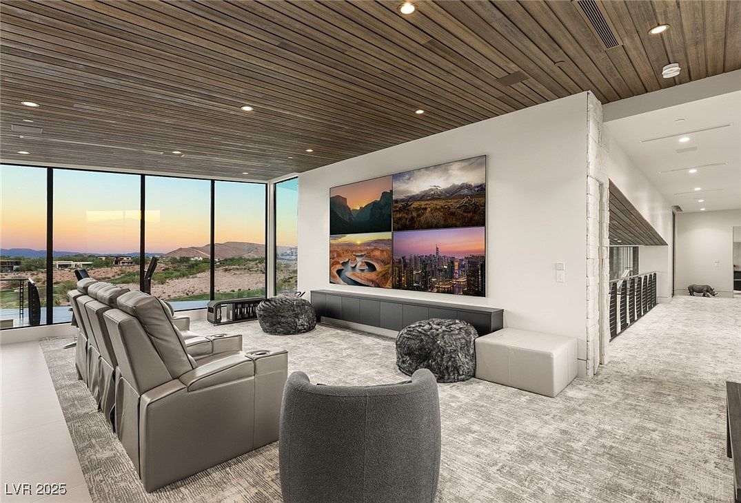 This is a modern living room featuring a large media display above a sleek console, comfortable seating including reclining chairs and unique poufs, and a wall of windows offering a scenic view. The room is decorated in neutral tones with a textured rug and a wood-paneled ceiling, creating a sophisticated and inviting atmosphere. The perspective is from the back of the room, looking towards the window.