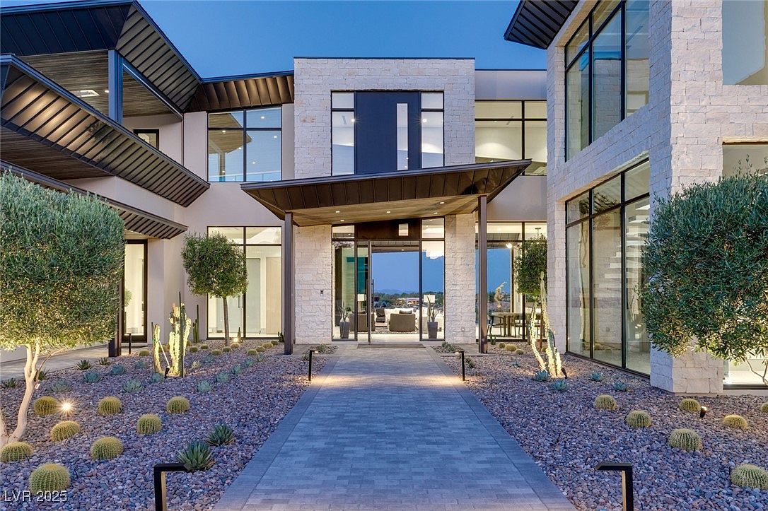 This is a stunning exterior shot of a modern home's entryway, featuring a stone facade, large windows, and a covered entrance. The walkway is paved with gray stone, leading to a glass front door, and the landscaping includes desert plants and subtle lighting, creating an inviting and luxurious feel. The architecture is contemporary with clean lines and a sophisticated color palette.