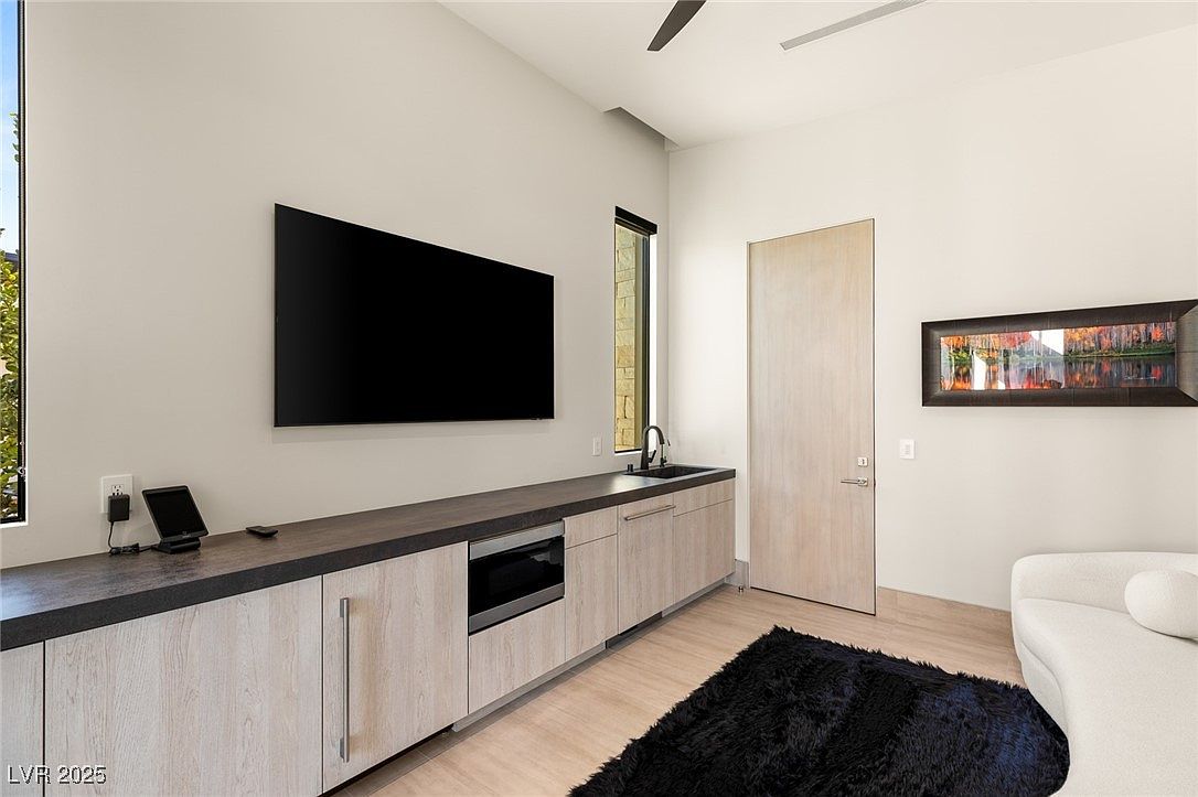 This interior shot showcases a modern living room area with a sleek entertainment center featuring a large flat-screen TV, built-in cabinets, and a sink. A contemporary artwork hangs on the wall, complemented by a minimalist white sofa and a plush black rug, creating a stylish and comfortable atmosphere. The room is well-lit, highlighting the clean lines and modern design elements.