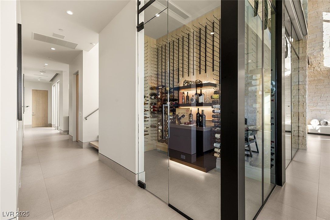 This interior shot showcases a modern hallway leading to a wine cellar. The hallway features neutral-toned tile flooring, recessed lighting, and clean white walls. The wine cellar is enclosed by glass, displaying an extensive wine collection and a sleek, contemporary design, adding a luxurious touch to the home.