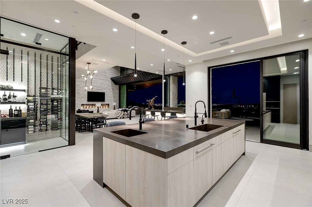 This is a modern, high-end kitchen featuring a large island with a dark countertop and light wood cabinetry. The kitchen includes a wine cellar, and opens to a living area with a fireplace and a view of the outdoors through large sliding glass doors. The space is well-lit with recessed lighting and pendant lights, creating a luxurious and inviting atmosphere.