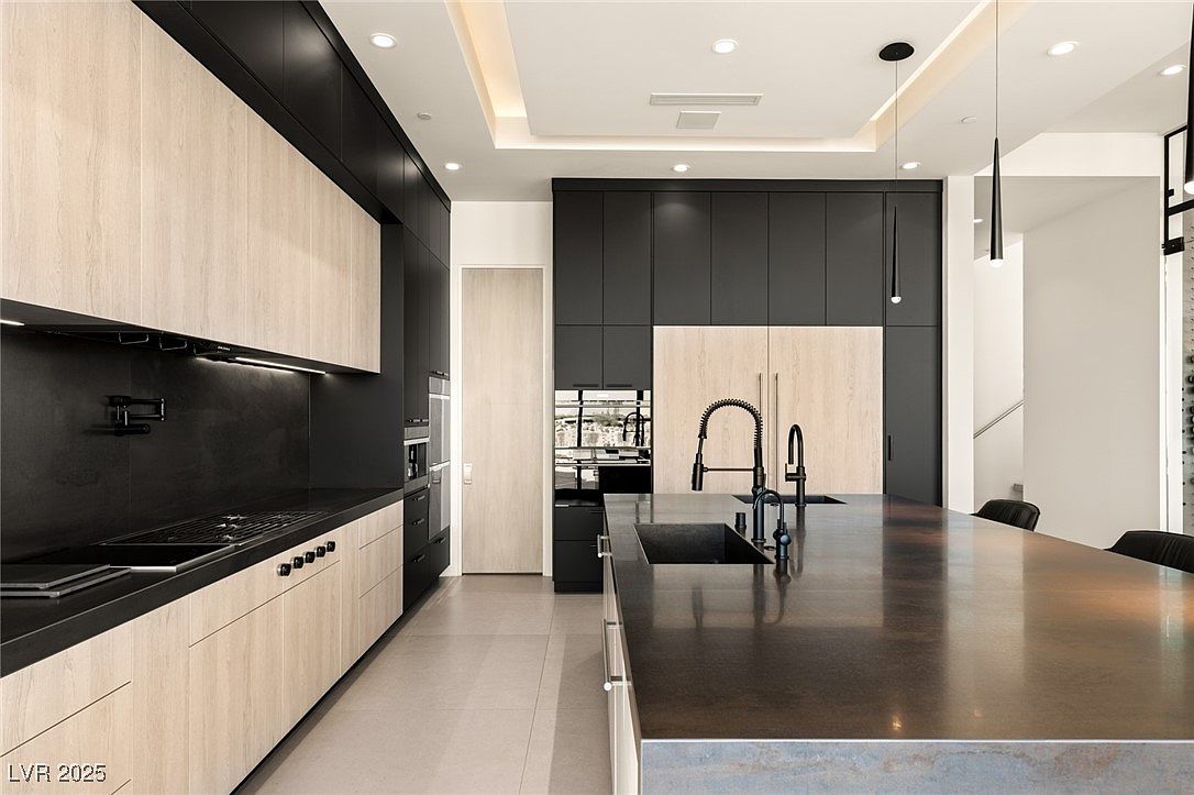 This is a modern kitchen with a large island featuring a dark countertop and dual sinks. The cabinets are a mix of light wood and dark finishes, creating a sleek and contemporary look. Stainless steel appliances are integrated into the cabinetry, and pendant lights hang above the island, adding to the sophisticated ambiance.