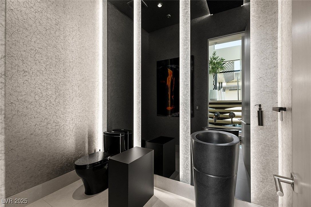 This is a modern guest bathroom featuring a black toilet, a black pedestal sink, and a sleek, minimalist design. The walls are covered in a textured wallpaper, and the lighting is provided by vertical light strips flanking a large mirror. The overall impression is one of sophistication and luxury.