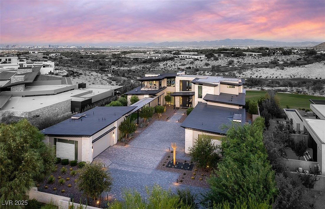 This aerial view showcases a modern luxury home with a flat roof, stone accents, and a spacious driveway. The property is surrounded by desert landscaping and offers panoramic views of the city skyline and mountains in the distance. The overall impression is one of exclusivity and high-end living.