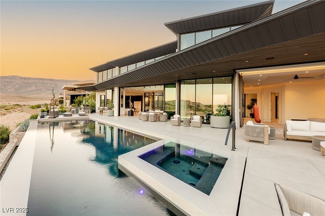 This image showcases a luxurious modern home's exterior, focusing on the pool and spa area. The infinity pool seamlessly blends with the horizon, while a separate spa with glass panels adds a touch of elegance. The expansive patio features contemporary outdoor furniture, creating an inviting space for relaxation and entertainment, all set against a backdrop of desert landscape and a stunning sunset.