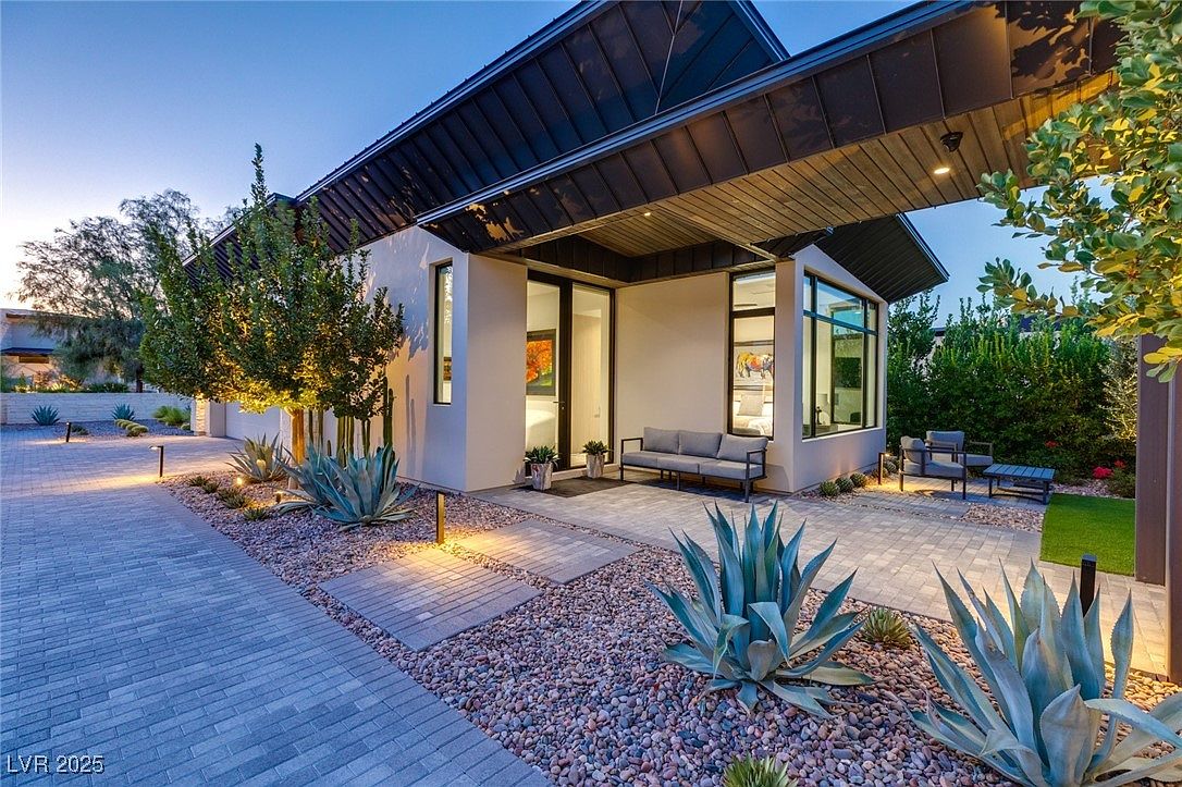 This image showcases the front exterior of a modern home with a striking architectural design. The property features a dark-colored roof, light-colored stucco walls, and large windows that provide ample natural light. The landscaping includes desert-style plants and decorative gravel, enhancing the home's curb appeal and creating a welcoming entrance.