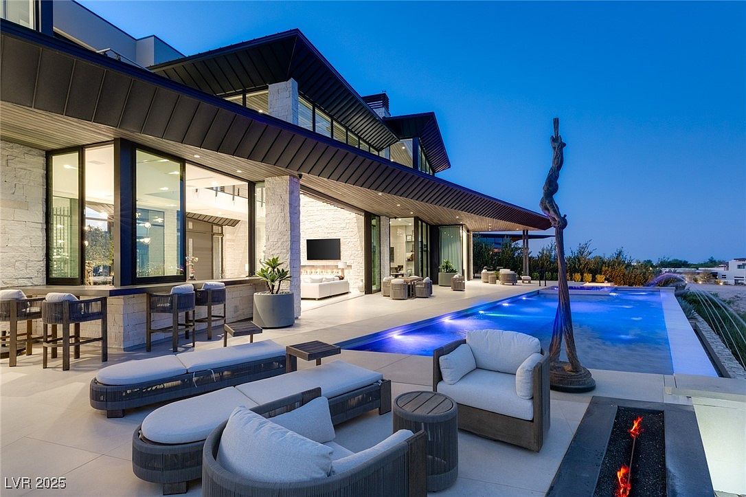 This image showcases a luxurious outdoor living space featuring a modern home with expansive windows and a sleek, dark roofline. The patio is furnished with stylish lounge chairs and seating areas, leading to a stunning pool with integrated lighting. A unique sculptural element stands beside the pool, adding an artistic touch, while a fire pit provides warmth and ambiance.