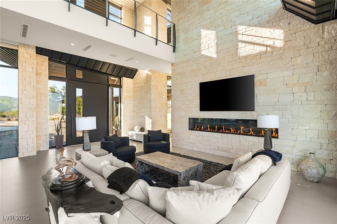 This is a spacious living room featuring a stone accent wall with a linear fireplace and a large television. The room is furnished with a large sectional sofa, armchairs, and a unique coffee table. The high ceilings and large windows create a bright and airy atmosphere, enhancing the modern design.