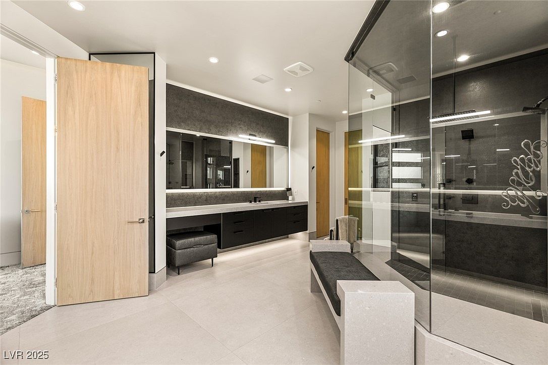 This is a modern primary bathroom featuring a double vanity with dark cabinetry and a light countertop, a large mirror with integrated lighting, and a glass-enclosed shower. A bench is placed near the shower, and the flooring is light-colored tile. The overall impression is luxurious and contemporary.