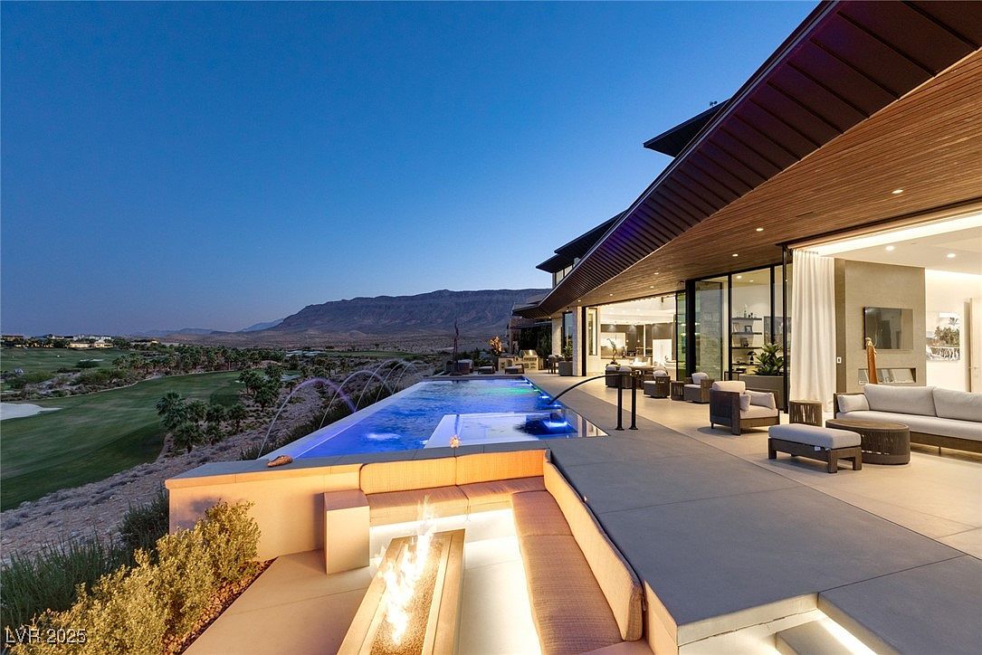 This image showcases a luxurious outdoor living space with a stunning infinity pool overlooking a golf course and mountain range. The area features a modern fire pit surrounded by built-in seating, a spacious patio with contemporary furniture, and seamless indoor-outdoor transition through large glass doors. The overall impression is one of high-end relaxation and sophisticated design.