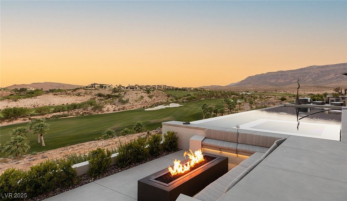 This image showcases a luxurious outdoor patio area with a modern fire pit and built-in seating. The patio overlooks a beautifully landscaped golf course and distant mountains, creating a serene and upscale ambiance. An infinity pool adds to the property's appeal, suggesting a high-end, resort-style living experience.