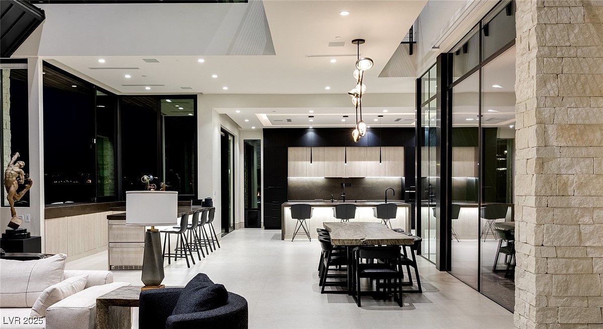 This interior shot showcases a modern living space that seamlessly blends with the kitchen and dining area. The open floor plan features sleek, minimalist design elements, including a stone accent wall, contemporary lighting fixtures, and large windows offering a view of the city lights. The color palette is neutral, creating a sophisticated and inviting atmosphere perfect for entertaining or relaxing.