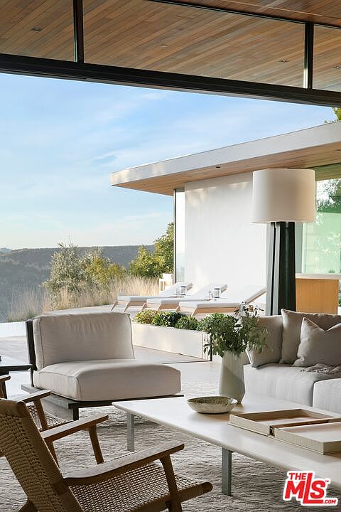 This sophisticated outdoor living space seamlessly blends indoor comfort with breathtaking natural views, featuring a contemporary lounge area with plush white seating and a woven wood chair. The open-air design is framed by a sleek, modern architectural overhang and leads directly to a sun-drenched terrace with lounge chairs overlooking a scenic hillside. The overall aesthetic is one of refined luxury, emphasizing a tranquil, high-end lifestyle perfect for relaxation and entertaining.