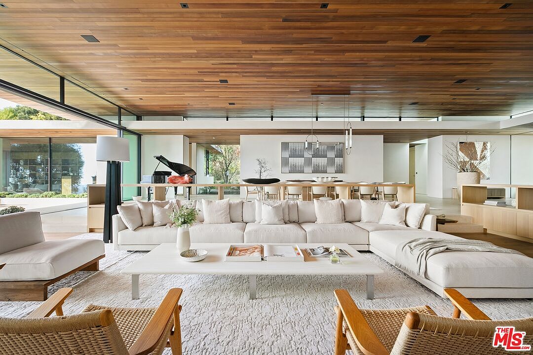 This expansive, open-concept living room showcases a sophisticated modern aesthetic with a warm wood-paneled ceiling and floor-to-ceiling glass walls that invite natural light. The space is anchored by a large, plush cream-colored sectional sofa and a minimalist white coffee table, complemented by woven wooden armchairs. In the background, a dining area and a grand piano are visible, emphasizing the home's airy, luxurious, and seamless indoor-outdoor flow.