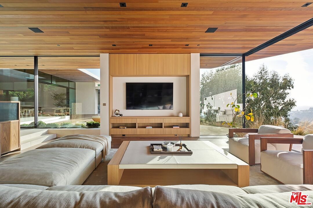 This sophisticated, modern living room features a warm wood-paneled ceiling and floor-to-ceiling glass walls that seamlessly blend the indoor space with the natural landscape outside. The room is anchored by a custom light-wood media console with a mounted television, complemented by a large, low-profile coffee table and plush, neutral-toned seating. The open-concept design creates an airy, tranquil atmosphere, emphasizing luxury and a strong connection to the outdoors.