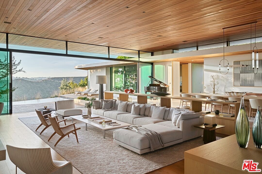 This expansive, open-concept living room features a seamless indoor-outdoor flow with floor-to-ceiling glass walls that reveal a stunning hillside vista. The space is anchored by a large, neutral-toned sectional sofa and mid-century modern accent chairs, all set beneath a warm, wood-paneled ceiling. A grand piano sits elegantly in the background, contributing to the sophisticated, airy, and luxurious atmosphere of this contemporary home.