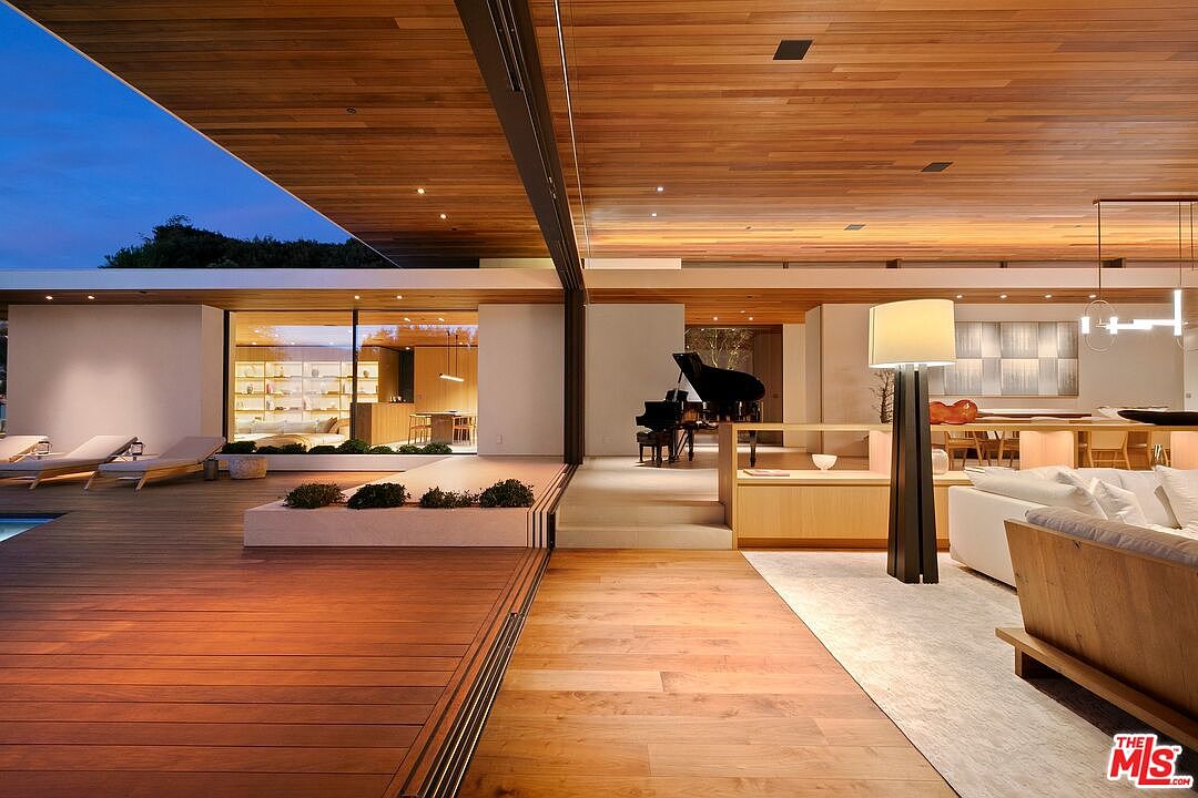 This sophisticated open-concept living space seamlessly blends indoor and outdoor living through expansive floor-to-ceiling glass walls that open onto a spacious wooden deck. The interior features warm wood-paneled ceilings, a grand piano, and a minimalist aesthetic, creating a luxurious and tranquil atmosphere. The perspective captures the transition from the polished indoor flooring to the exterior deck, highlighting the architectural harmony and high-end design of the residence.