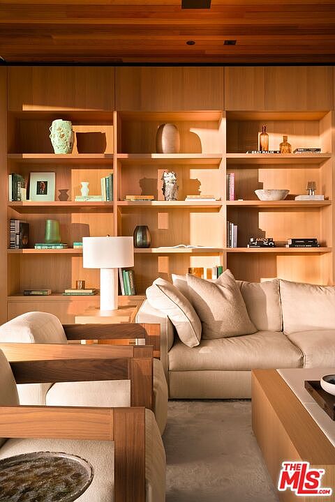 This sophisticated living room features a warm, natural wood-paneled wall with integrated floor-to-ceiling shelving, showcasing curated decor and books. A plush, neutral-toned sofa and a modern wooden armchair create a cozy seating area, complemented by a minimalist table lamp. The space is bathed in soft, golden natural light, emphasizing the high-end, contemporary aesthetic and inviting atmosphere.