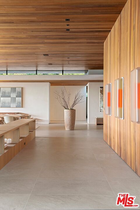 This spacious, minimalist hallway features a striking wood-paneled ceiling and a matching accent wall adorned with modern, illuminated orange-toned wall sconces. The open floor plan leads the eye toward a central decorative vase with branches, creating a serene and sophisticated atmosphere. Large-format light-colored floor tiles and clean white walls enhance the airy, architectural aesthetic of this contemporary home.