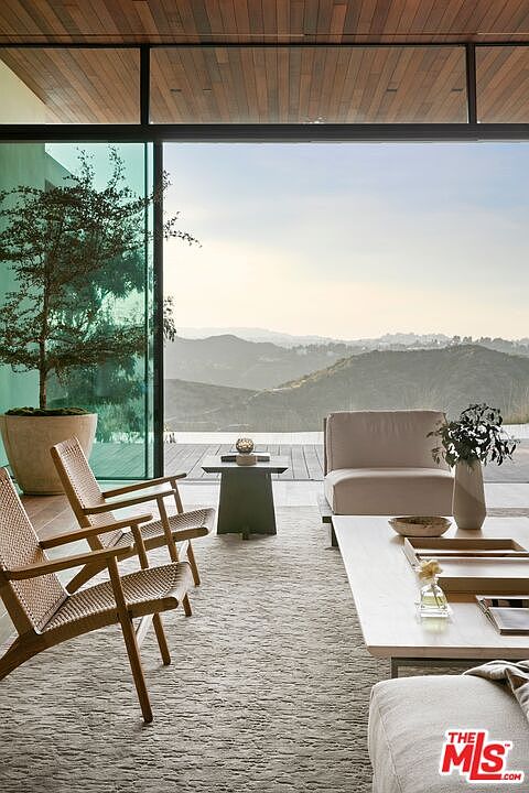 This sophisticated indoor-outdoor living space features a minimalist design with floor-to-ceiling glass walls that frame a breathtaking panoramic view of rolling hills. The room is furnished with elegant, light-toned woven chairs and a plush sofa, centered around a sleek coffee table on a textured area rug. The warm wood-paneled ceiling and seamless transition to the outdoor deck create an airy, serene atmosphere perfect for luxury living.