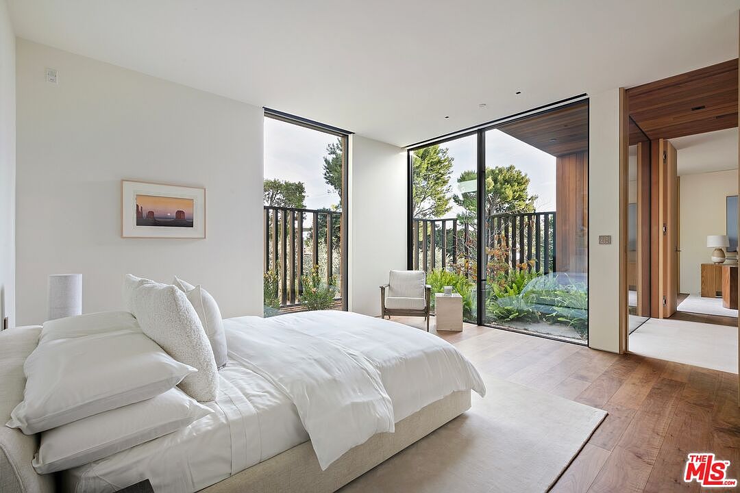 This serene primary bedroom features a minimalist, modern aesthetic with a plush white bed as the focal point. Floor-to-ceiling glass walls provide an expansive view of the lush, private garden outside, seamlessly blending indoor and outdoor living. The room is finished with warm hardwood flooring and clean white walls, creating a tranquil and sophisticated atmosphere.