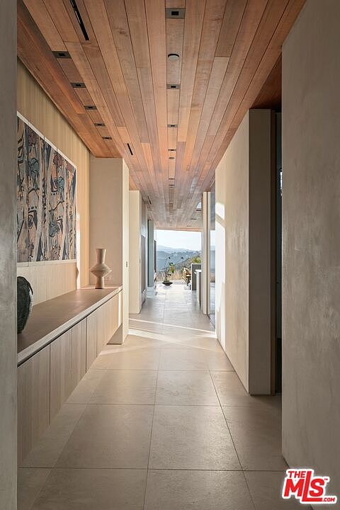 This elegant, minimalist hallway features a striking wood-paneled ceiling with recessed lighting and large-format light-toned floor tiles. A long, custom built-in console provides storage on the left, complemented by abstract wall art, while the perspective draws the eye toward a bright, open doorway revealing a scenic outdoor view. The space exudes a sophisticated, modern aesthetic with clean lines and a seamless transition between indoor and outdoor living.