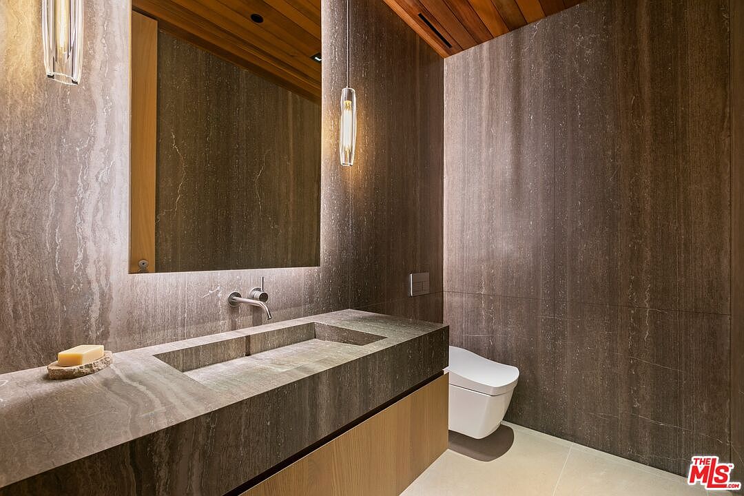This sophisticated powder room features a striking stone-clad wall and a custom integrated stone sink, creating a luxurious, minimalist aesthetic. The space is accented by warm wood ceiling paneling, sleek pendant lighting, and a modern wall-mounted toilet. The overall impression is one of high-end, contemporary design with a focus on natural textures and clean lines.