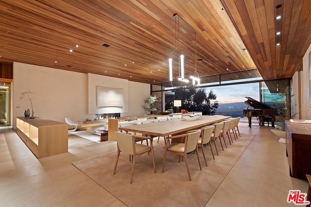 This expansive, open-concept dining room features a long wooden table surrounded by minimalist chairs, set beneath a striking modern chandelier. The room is defined by its warm wood-paneled ceiling, recessed lighting, and floor-to-ceiling glass walls that reveal a breathtaking sunset view over the landscape. A grand piano sits in the corner, while a sleek fireplace and neutral-toned living area are visible in the background, creating a sophisticated and luxurious atmosphere.