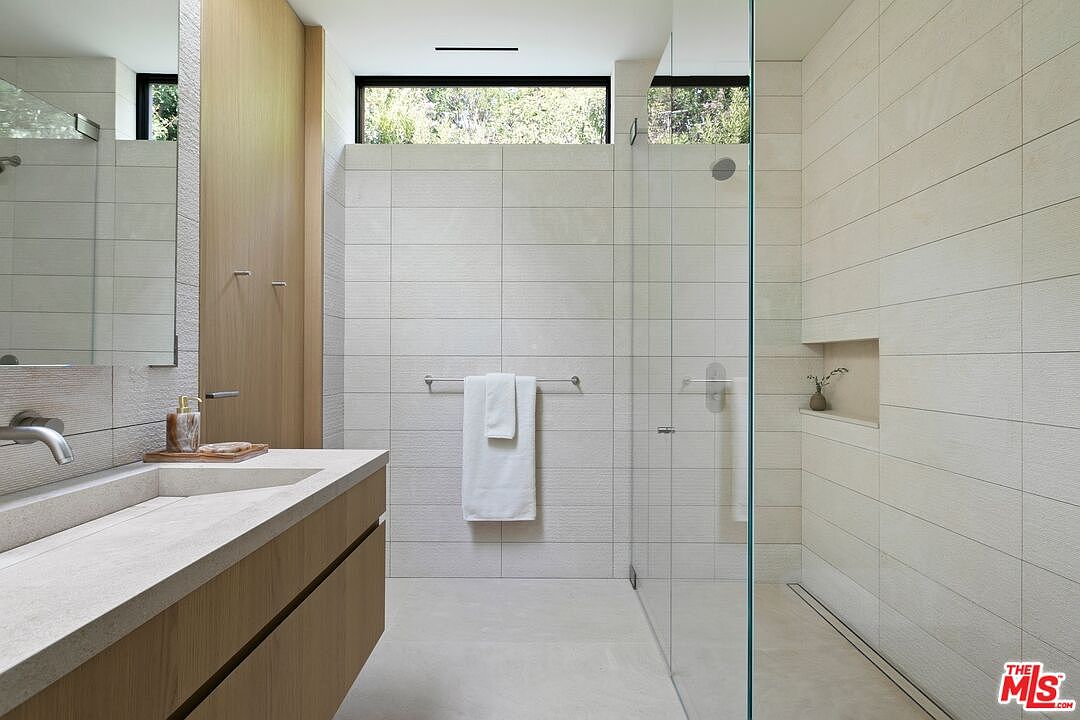 This modern, minimalist primary bathroom features a sleek floating vanity with a stone basin and light wood cabinetry. A spacious walk-in shower is enclosed by a frameless glass panel, showcasing textured wall tiles and a built-in recessed niche. The room is brightened by a high, horizontal clerestory window that allows natural light to filter into the space, creating a serene and spa-like atmosphere.