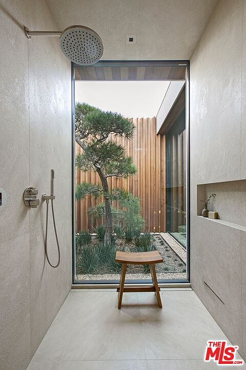 This luxurious, minimalist shower features large-format stone tiling and a high-end rainfall showerhead with a handheld attachment. A floor-to-ceiling glass wall provides a serene, direct view of a private courtyard garden with a sculptural pine tree and wooden slat fencing. A simple wooden stool sits in the center, enhancing the spa-like, tranquil atmosphere of this indoor-outdoor design.