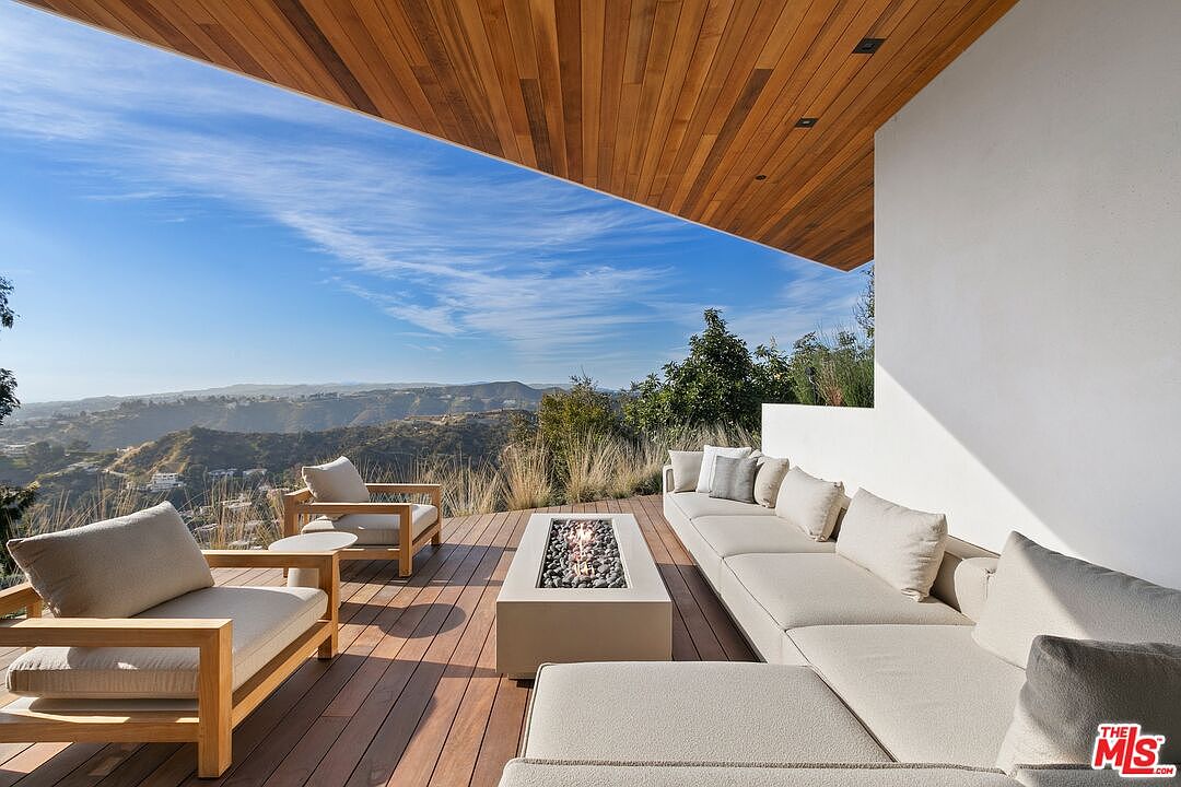 This stunning outdoor deck features a modern, minimalist design with a warm wood-paneled ceiling and expansive panoramic views of the surrounding hills. The space is furnished with a sleek, light-toned sectional sofa and matching armchairs arranged around a contemporary rectangular fire pit. The open-air layout creates a seamless transition between indoor luxury and the natural landscape, perfect for high-end entertaining.