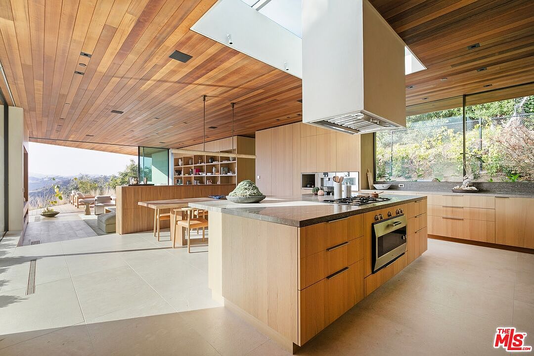 This stunning, modern open-concept kitchen features warm wood cabinetry, a large central island with a stone countertop, and a sleek stainless steel oven. The space is defined by a dramatic wood-paneled ceiling and floor-to-ceiling glass walls that seamlessly blend the interior with a spacious outdoor patio and scenic hillside views. The design emphasizes a minimalist, high-end aesthetic with abundant natural light pouring in from a large skylight above the range hood.