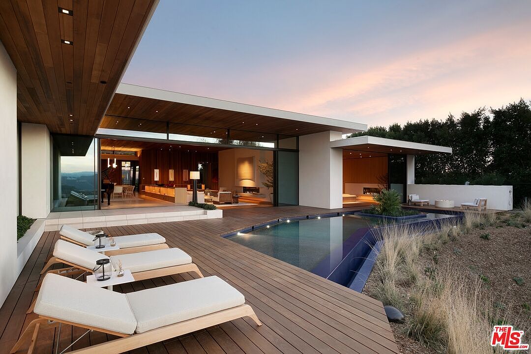 This stunning modern luxury home features a seamless indoor-outdoor flow, highlighted by a spacious wooden deck and a sleek, geometric infinity-edge pool. The open-concept living area is visible through expansive glass sliding doors, showcasing a sophisticated interior design at dusk. The scene is framed by natural landscaping and a warm, ambient sunset, creating an atmosphere of high-end tranquility and architectural elegance.