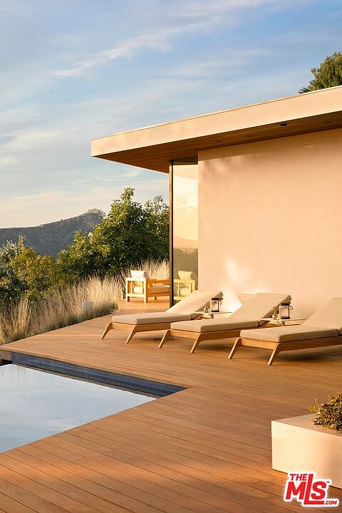 This stunning modern deck features a sleek infinity-edge pool overlooking a serene, mountainous landscape at sunset. The space is furnished with minimalist lounge chairs, creating a tranquil and luxurious outdoor living area. The warm, golden hour lighting enhances the clean architectural lines and natural wood decking, evoking a high-end, resort-like atmosphere.