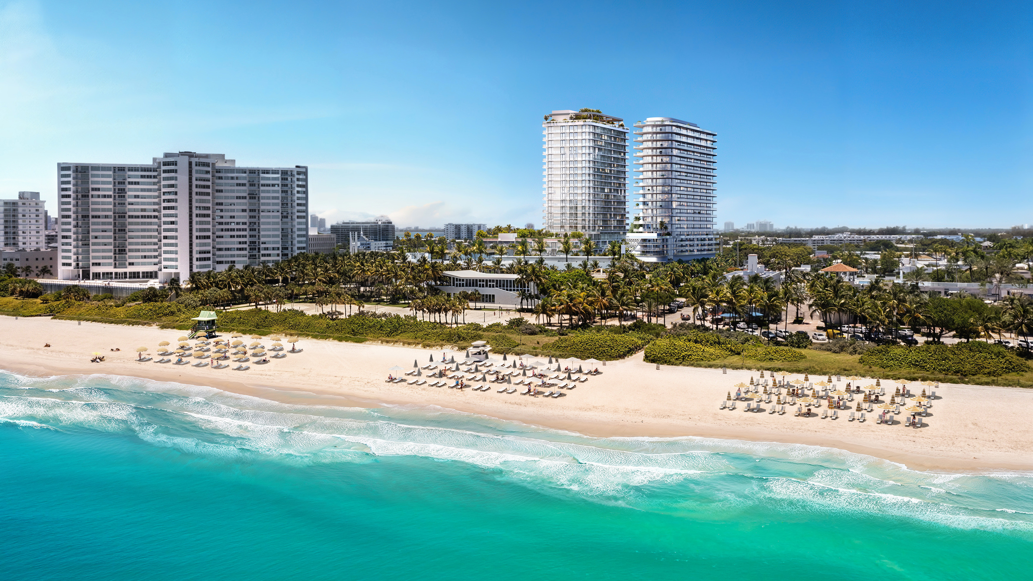 An aerial view showcases luxury beachfront property with modern high-rise buildings. The scene highlights a pristine beach with neatly arranged beach chairs and umbrellas, contrasted by the clear turquoise ocean. Palm trees and manicured landscapes contribute to the upscale, resort-like ambiance, making it an appealing listing for potential buyers seeking luxury coastal living.