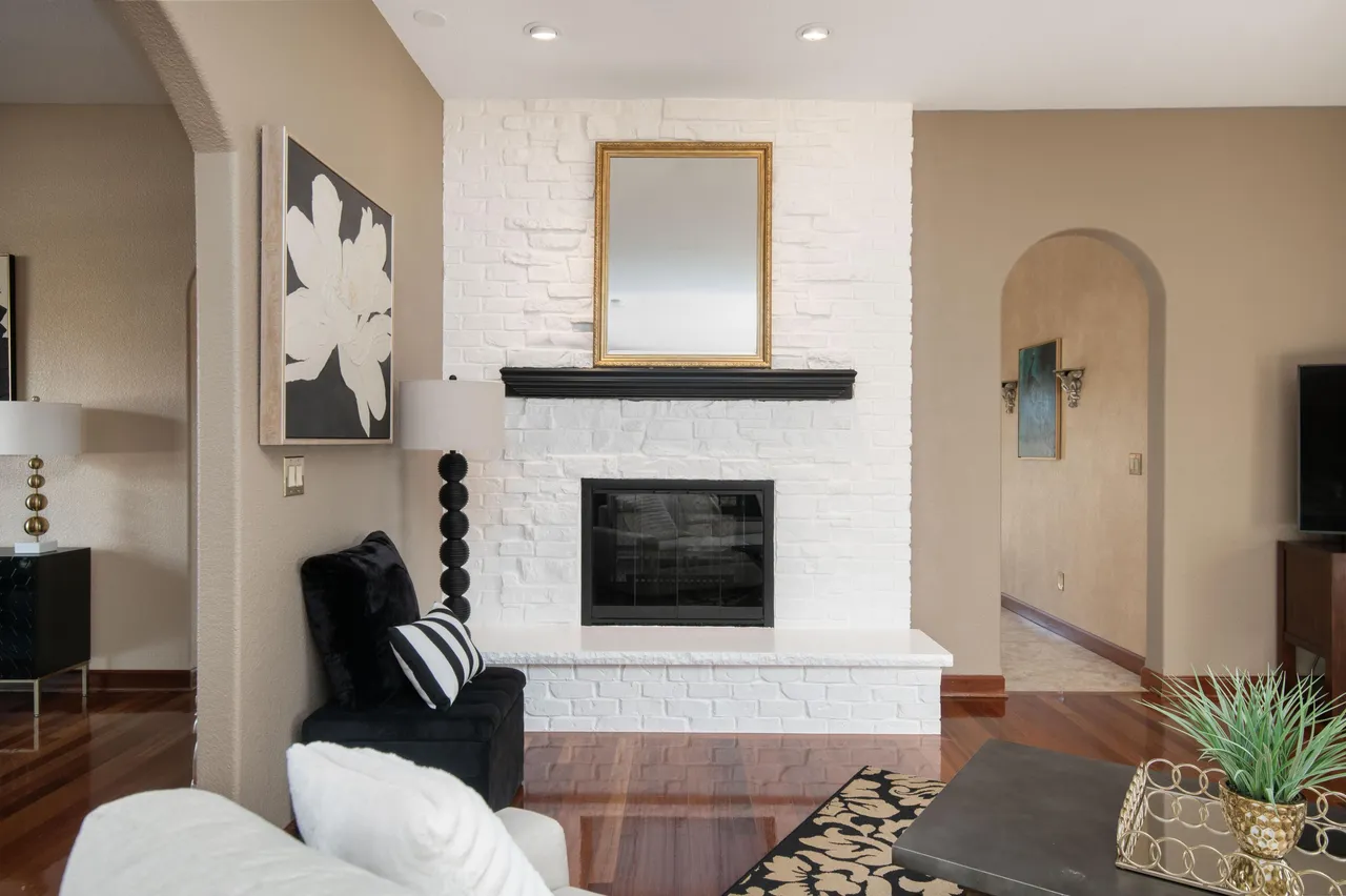 This living room features a white-painted brick fireplace with a black mantel and a gold-framed mirror hanging above. Comfortable seating includes a white sofa and a black velvet ottoman with a striped pillow. The room has hardwood floors and a neutral color palette, creating a warm and inviting atmosphere.
