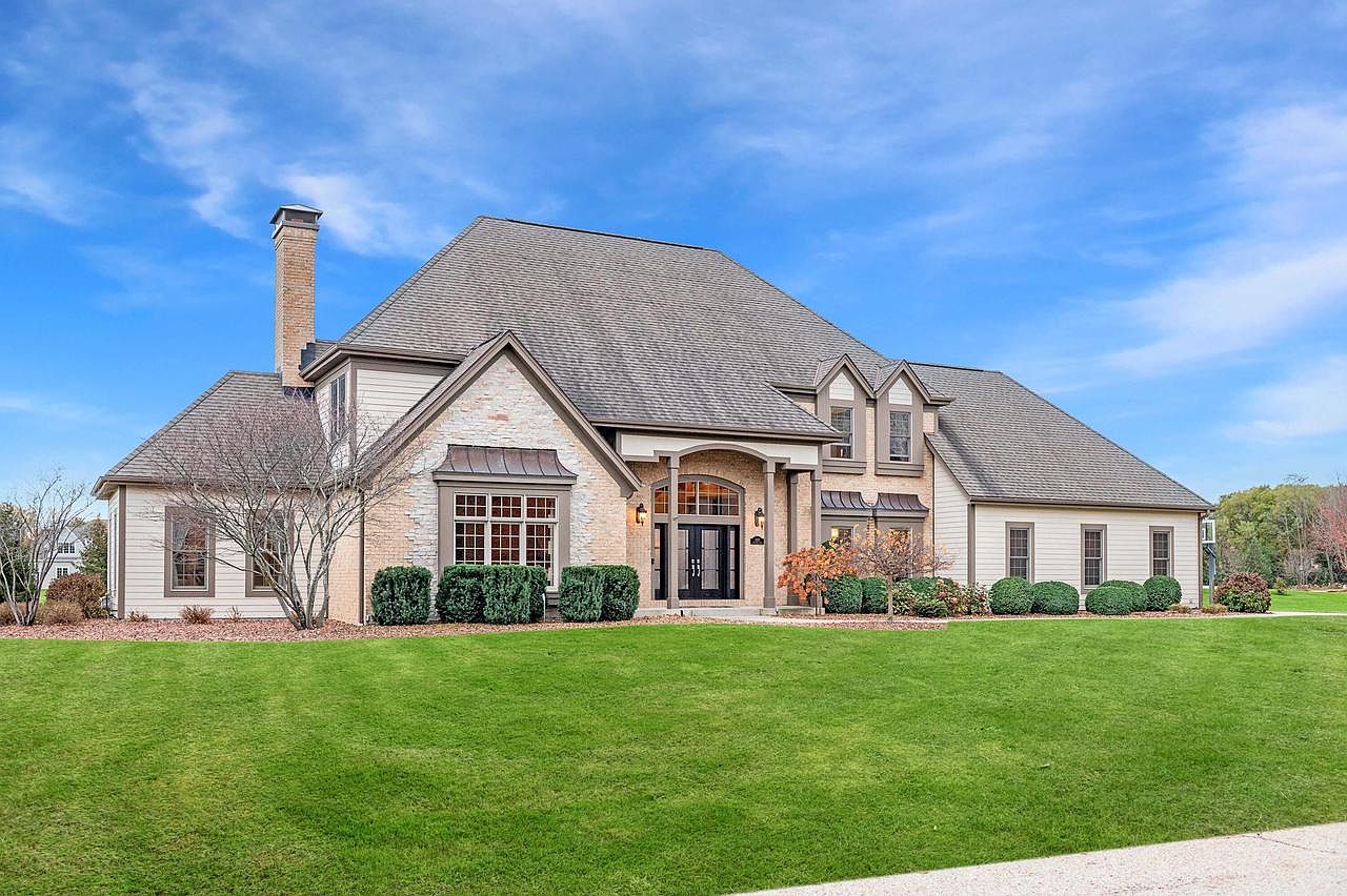 This is a front view of a large, luxurious two-story house with a well-manicured lawn. The house features a combination of brick and siding, a prominent roofline, and a grand entryway with double doors. The landscaping includes neatly trimmed bushes and trees, enhancing the property's curb appeal.