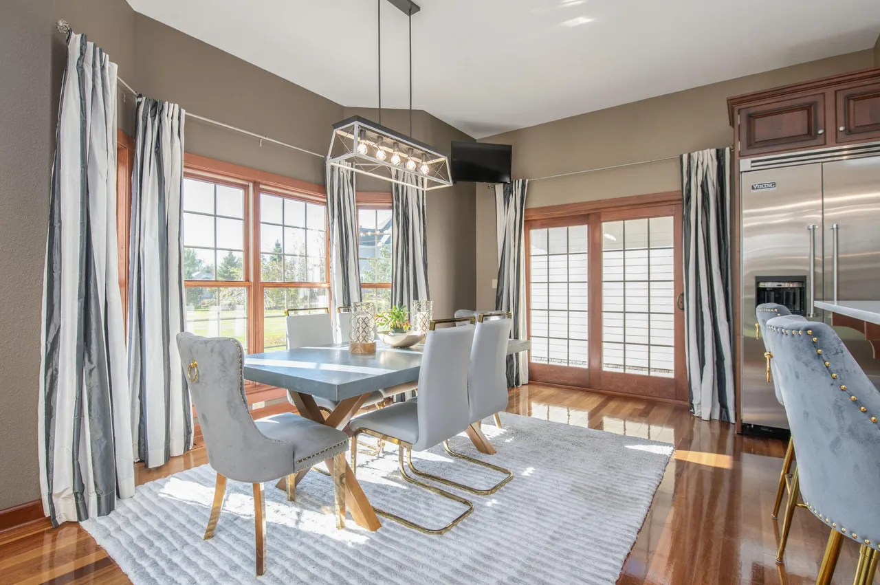 This is an interior shot of a dining room featuring a large grayish tabletop with wooden legs. There are white cushioned chairs surrounding the table. The flooring is wooden and covered by a white rug. Floor-to-ceiling black and white striped curtains drape from the windows.