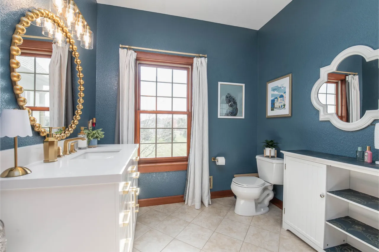 This is a charming guest bathroom with a classic design. The walls are painted a deep blue, complementing the beige tiled floor. The white vanity with gold fixtures and an oval mirror adds a touch of elegance, while the natural light from the window brightens the space.