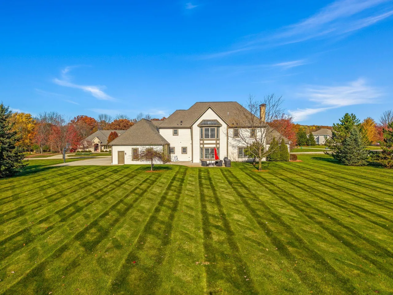 This is a well-manicured rear exterior view of a large house. The yard features expertly striped green grass leading up to the house's rear, detailing a cozy patio area with an umbrella and outdoor seating. The architecture combines traditional elements with modern conveniences, set against a clear blue sky with scattered clouds.