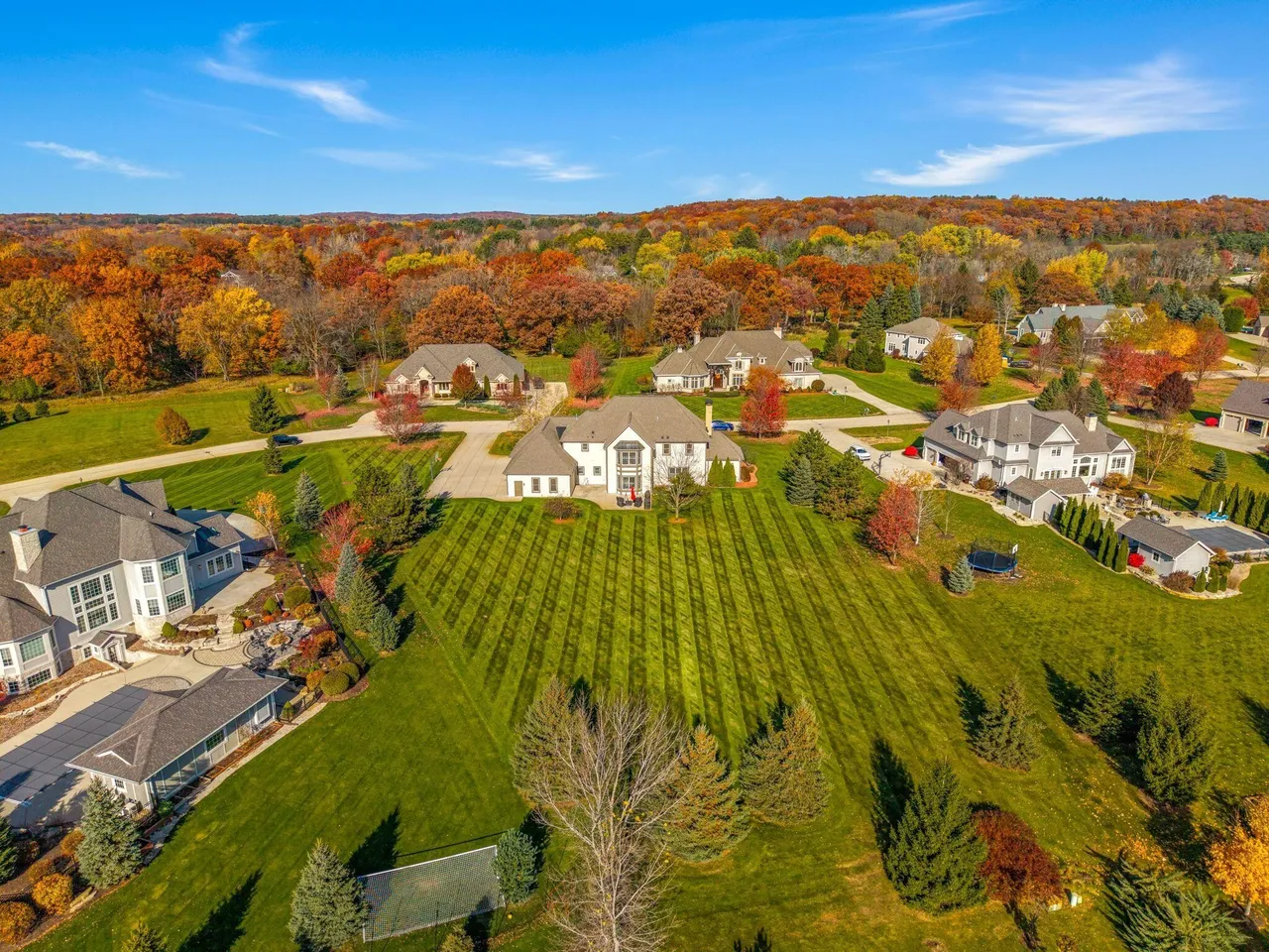 This aerial view showcases a stunning property featuring a large, well-maintained lawn with striped mowing patterns, complemented by mature trees displaying vibrant fall foliage. The house is a prominent feature, blending architectural design with the surrounding natural landscape. Several other upscale homes can be seen in the neighborhood, indicating a high-end residential area.