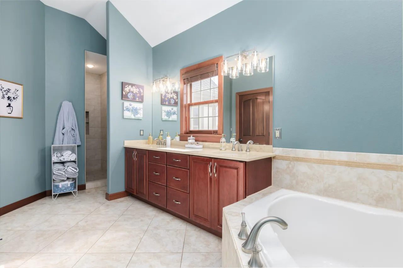 This is a primary bathroom featuring a double sink vanity with dark wood cabinetry and a light countertop. The walls are painted a calming blue, and there's a separate bathtub. The room also has a window and a well-lit mirror, creating a bright and functional space.