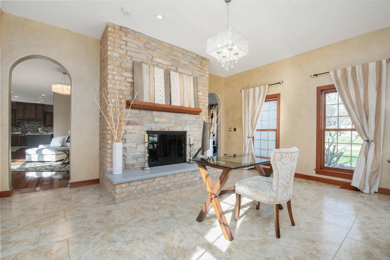 This dining room features a prominent brick fireplace with a wooden mantle, complemented by decorative vases and candles. A modern glass-topped table with a unique base is paired with an upholstered chair, adding a touch of elegance. The room is illuminated by a decorative chandelier and natural light streaming through the windows with striped curtains.