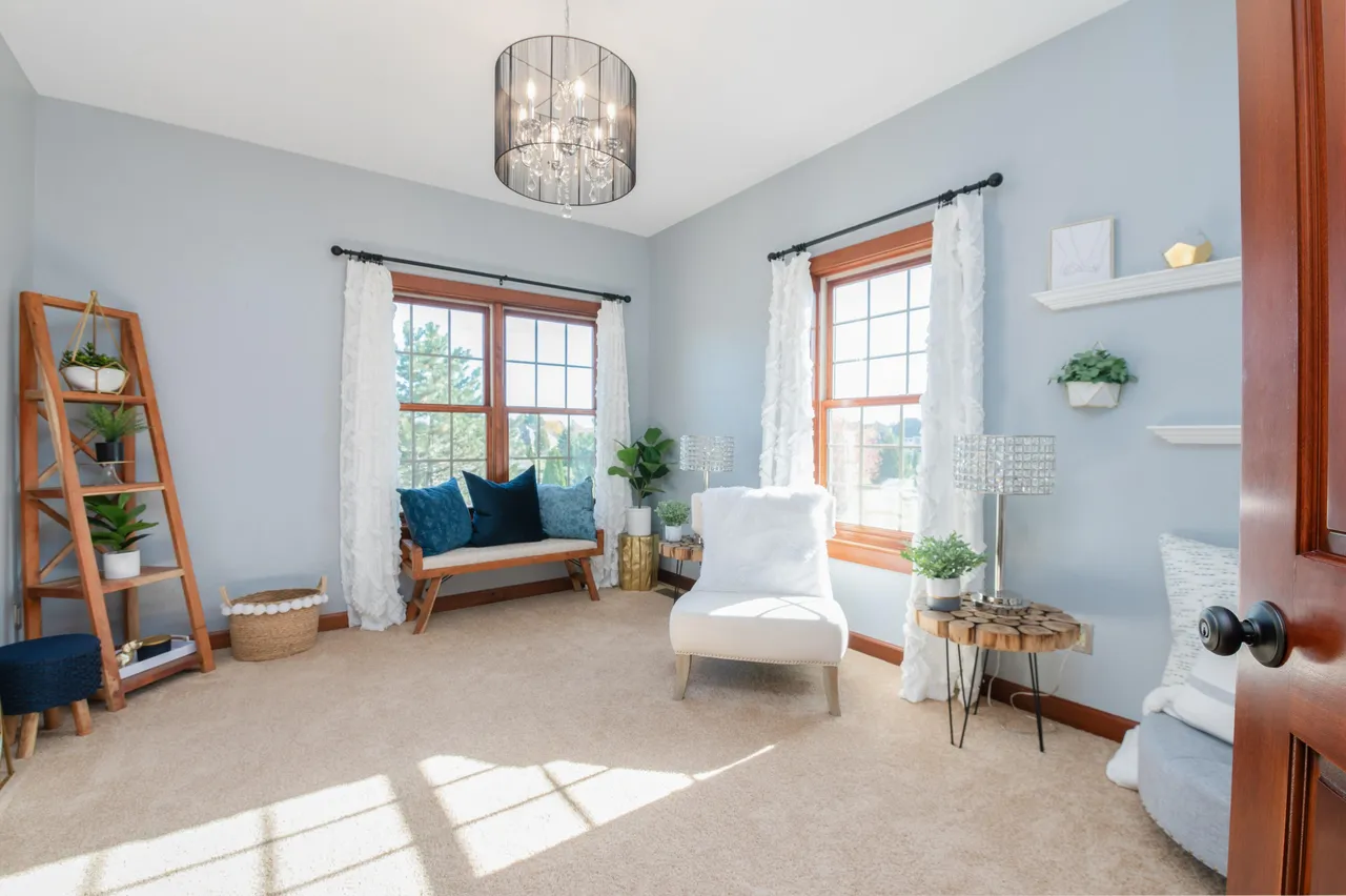 This is a staged guest bedroom with a neutral color scheme and natural light. Features include two windows with ruffled white curtains, a bench seat with blue pillows and a stylish light fixture. The room is furnished with a white armchair, a wood side table, and a decorative ladder. The overall impression is bright, cozy, and inviting.
