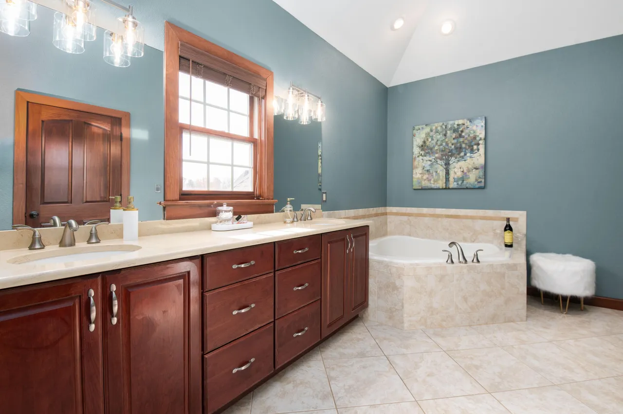 This primary bathroom features a double vanity with dark wood cabinetry and light countertops. A soaking tub with a tiled surround is adjacent to the vanity, and the walls are painted a calming blue hue. The tile flooring and window with a wooden frame add warmth to the space.