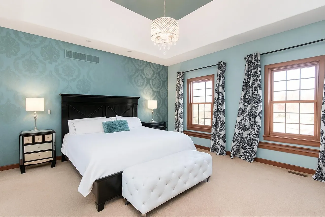 This is a primary bedroom featuring an elegant design with blue patterned wallpaper and two large windows framed by patterned curtains. The focal point is a king-sized bed with a dark headboard, crisp white linens, and a tufted white bench. The room is well-lit and appears spacious, contributing to a luxurious and comfortable ambiance.