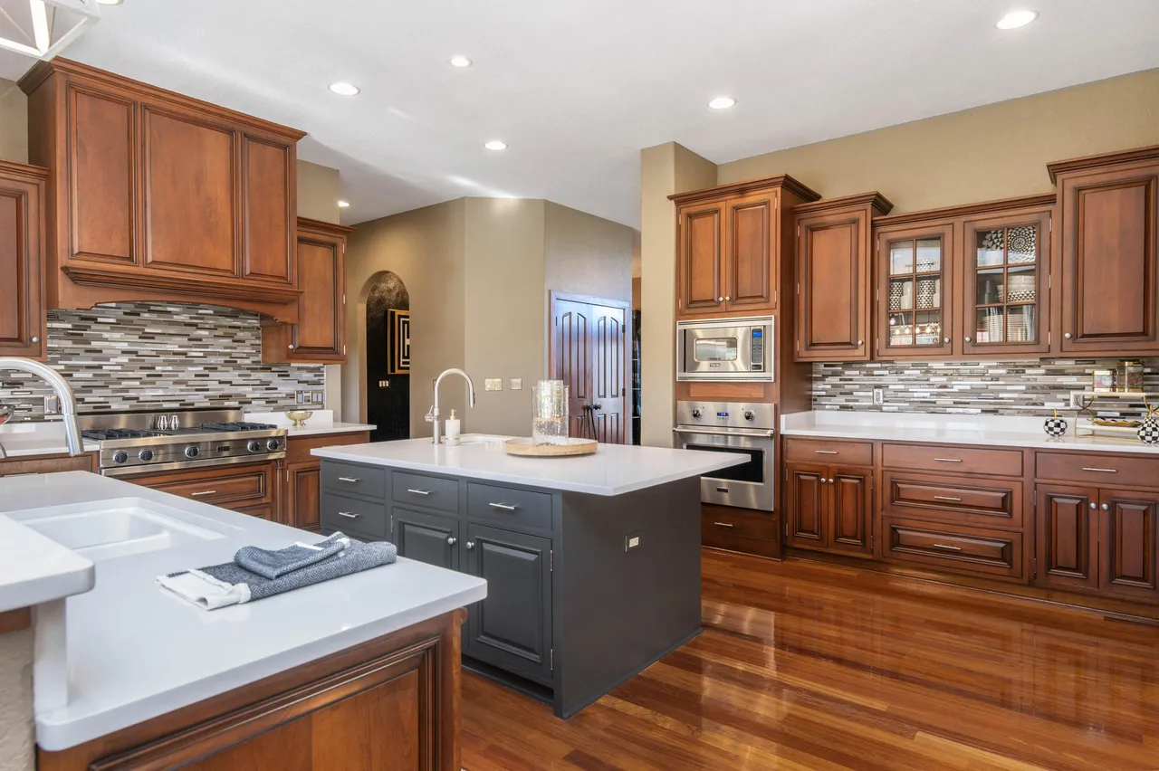 The image showcases a well-appointed kitchen with rich, wood cabinetry and a contrasting dark gray island. The kitchen features stainless steel appliances and a mosaic backsplash, lending a touch of modern style to the traditional design. Hardwood floors reflect ample lighting, contributing to a warm and inviting atmosphere.