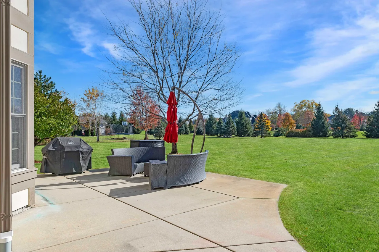 This image showcases a concrete patio area in a backyard setting. The patio features outdoor furniture, including a curved sectional sofa and a grill covered with a protective tarp. The yard is well-maintained with lush green grass and landscaping, suggesting a comfortable outdoor living space.